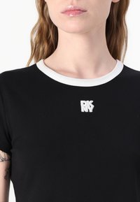 Čierne tričko s bielym rebrovaným golierom a vyšitým logom "DKNY" na hrudi. Hladká textúra a krátke rukávy.