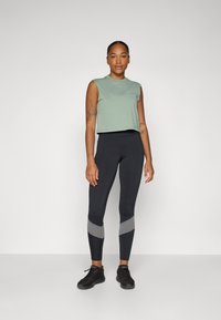 Green cropped tank top with sleeveless design paired with black leggings featuring gray accent stripes and black athletic shoes.