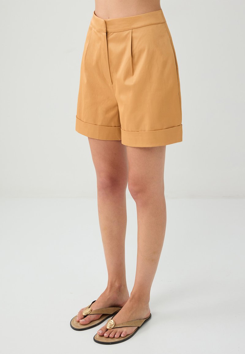 Shorts plissés beiges avec un ourlet retroussé, fabriqués en tissu lisse. Associés à des sandales plates accentuées de doré, mettant en valeur un look décontracté.