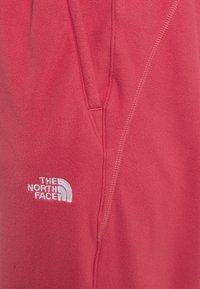 Tecido de fleece vermelho, apresentando um design costurado com um pequeno logótipo branco da "The North Face" no canto inferior esquerdo. Textura suave, sem padrões visíveis.