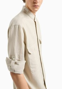 La camicia di lino beige presenta un colletto a stand, maniche arrotolate e due tasche sul petto con dettagli in bottoni. Texture liscia e leggera.