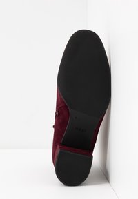 Bottine en daim bordeaux pour femme avec semelle en caoutchouc noire et petit talon bloc, vue de la semelle appuyée contre un coin blanc.