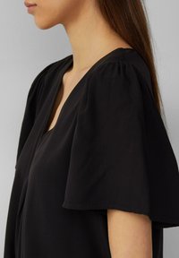 Blouse noire avec un décolleté en V, dotée de manches courtes bouffantes. Le tissu semble doux et léger, avec une texture lisse.