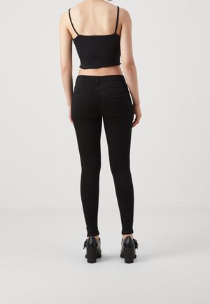 Vrouw draagt een zwart cropped tanktop, zwarte skinny jeans en zwarte schoenen met hak, staand met de rug naar voren op een effen achtergrond.