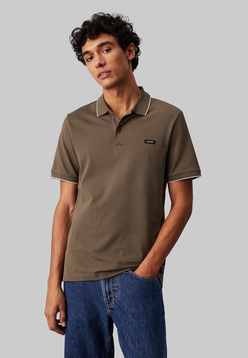 Polo shirt verde oliva con bordi bianchi sul colletto e sulle maniche, dotato di una piccola toppa con logo sul petto sinistro. Maniche corte.