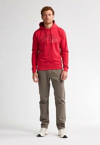 Felpa rossa con cappuccio e tasca a marsupio, con testo grigio stampato sul petto. Abbinata a pantaloni kaki chiari e sneakers bianche. Texture liscia.