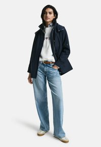 Marinblå vattentät jacka med hög krage och dragkedjor, parat med en vit sweatshirt, ljusblå vida jeans och beige sneakers.