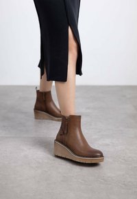 Botas de cuero marrón hasta el tobillo con cremallera lateral, suela de cuña y detalles texturizados. Combinadas con una falda negra con aberturas.