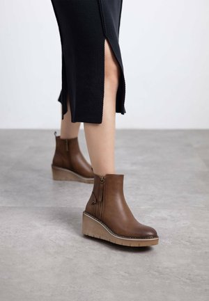 Botas de cuero marrón hasta el tobillo con cremallera lateral, suela de cuña y detalles texturizados. Combinadas con una falda negra con aberturas.