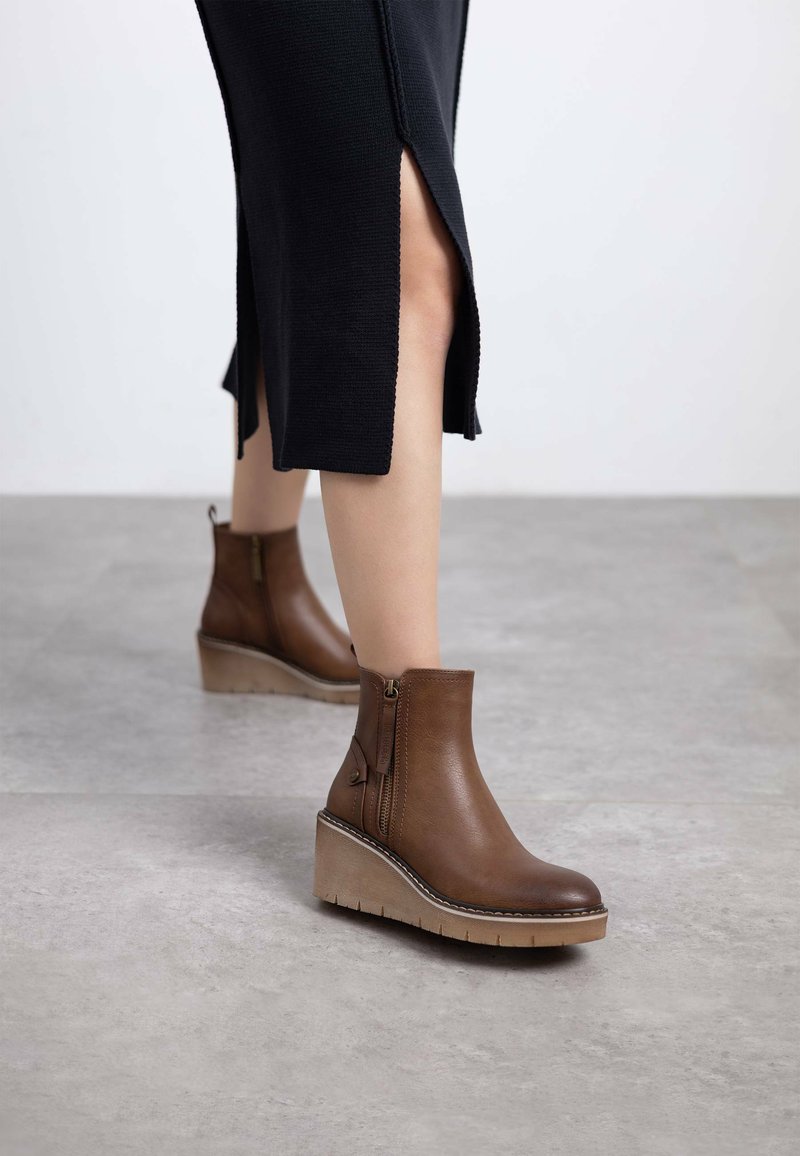 Botas de cuero marrón hasta el tobillo con cremallera lateral, suela de cuña y detalles texturizados. Combinadas con una falda negra con aberturas.