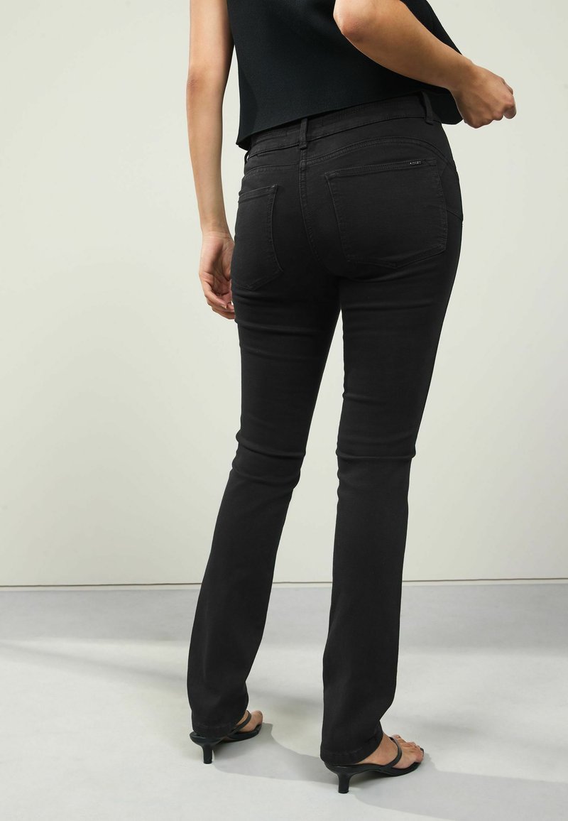 Jeans de mezclilla negra con corte ajustado, cintura alta y sutiles bolsillos traseros. Usados con tacones negros de punta abierta sobre un fondo neutro.