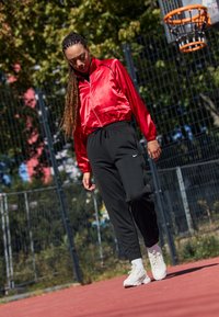 Sarkans satīna jaku ar rāvējslēdzēju, melnas sporta bikses ar elastīgu jostasvietu un baltas sporta kurpes. Brīvdabas basketbola laukuma fons.