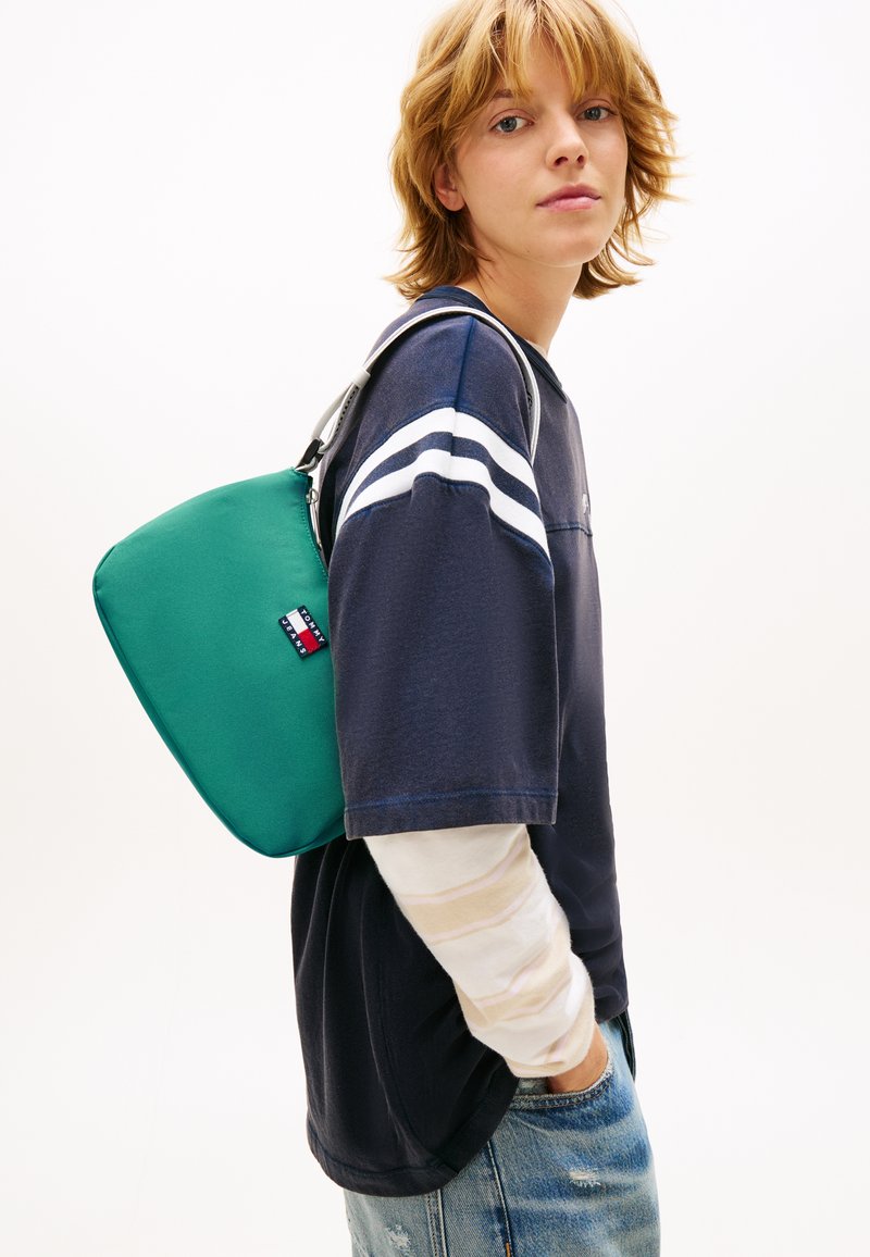 Πράσινη τσάντα ώμου με υφή στην επιφάν εια, με αντιθετικό λογότυπο, συνδυασμένη με μπλε σκούρο πουκάμισο και μανίκι με ανοιχτόχρωμες λεπτομέρειες από κάτω.