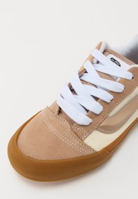 Baskets en suede beige avec des lacets blancs, des panneaux en tissu crème et une semelle en caoutchouc marron. Couture visible et étiquette logo sur la languette.