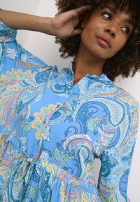 Svetlo modra obleka s patternom, ki vsebuje paisley vzorce v rumeni, zeleni in roza barvi. Gumbasta zapenjanje in vrvica za pas. Mehka tekstura blaga.