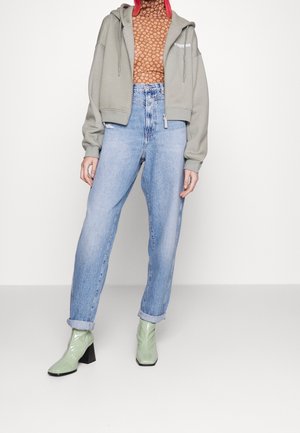 Grå kort hoodie med dragsko, brun mönstrad topp, högmidjade ljusblå jeans och glänsande gröna ankelboots med blockklackar.