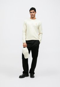 Long-sleeve cream shirt with a subtle logo, paired with black cargo pants and black shoes, holding a light gray cap. Simple design.