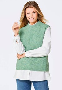 Grüner gerippter Strickpullover über einem weißen Hemd mit Kragen, mit hohem Ausschnitt und ärmellosem Design, kombiniert mit blauen Jeans.