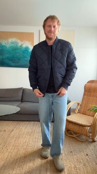 Casaco bomber preto acolchoado com acolchoamento em diamante, usado sobre uma camisa preta, combinado com jeans azul claro e sapatos cinzentos.