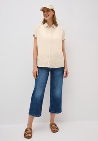 Beige overhemd met korte mouwen, blauwe wijde jeans, en bruine sandalen met studs; model draagt een lichtgekleurde pet.