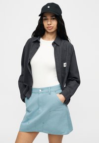 Νεαρή γυναίκα με μαύρο καπέλο, σκούρο πουκάμισο με κουμπιά πάνω από λευκό τοπ και ανοιχτό μπλε φούστα, με τα χέρια στις τσέπες, μπροστά σε απλό φόντο.