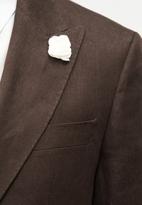 Καφέ σακάκι με ένα λευκό υφασμάτινο λουλούδι καρφιτσωμένο στην αριστερή πέτα και λευκό γιακά πουκάμισου ορατό από κάτω.