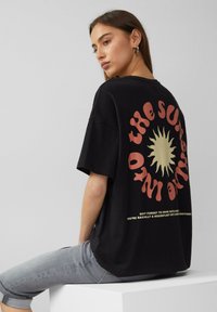 Schwarzes Baumwoll-T-Shirt mit einem großen Sonnen-Graphic und rotem und goldenem Text auf dem Rücken. Lässige, lockere Passform mit kurzen Ärmeln.