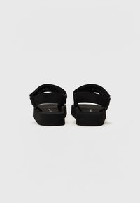 Des sandales noires avec une semelle plate et des sangles épaisses et texturées. Elles sont dotées de boucles ajustables et d'un design simple. La vue arrière met en avant la hauteur du talon.
