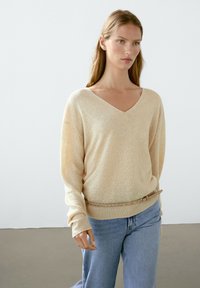 Beige gebreide trui met een V-hals, een ontspannen pasvorm en lange mouwen. Gecombineerd met lichtblauwe jeans en een bruine riem met metalen details.