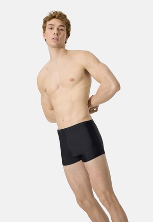 Joven sin camiseta con cabello rizado, vistiendo bañador negro, de pie ligeramente de lado con las manos detrás de la espalda sobre un fondo liso.