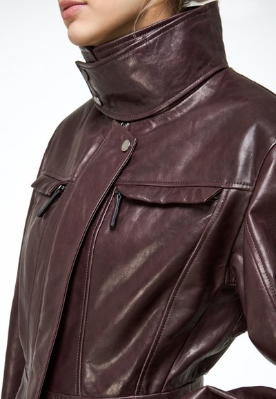 Femme portant une veste en cuir marron foncé avec un col montant, des poches zippées et des fermetures à boutons-pression.