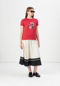 A woman stands in a red graphic tee featuring cartoon pets, paired with a white pleated skirt and black shoes, wearing sunglasses.