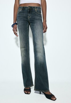 Femme portant un jean bootcut bleu délavé, des talons noirs ouverts, un top court blanc et un bracelet bleu foncé, debout devant un fond uni.