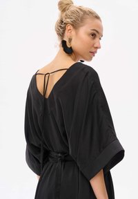 Schwarzes Kimono-Kleid mit V-Ausschnitt im Rücken, weiten Ärmeln und einer Bindeschließe in der Taille. Hergestellt aus einem glatten, leichten Stoff.