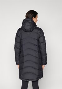 Svart puffjacka med huva, med ett chevron-quiltat mönster och en logga på baksidan. Slät textur och knälång design.