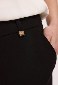 Czarne spodnie z złotym, kwadratowym akcentem sprzętowym z logo "M", textured tkanina oraz z pętelką na pasek w talii, częściowo zasłonięte.