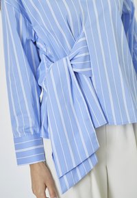Light blue shirt with white stripes, featuring a front knot detail and long sleeves. Fabric appears smooth and slightly glossy.