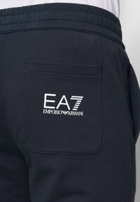 Tmavě modré tepláky s elastickým pasem, s jednou zadní kapsou. Na kapse je bílý potisk loga "EA7 EMPORIO ARMANI". Měkká látka, sportovní design.