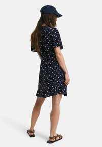 Marineblauwe polka dot jurk met korte mouwen, gerimpelde zoom en een verstelbare tailleband; gecombineerd met bruine platte sandalen en een bijpassende pet.