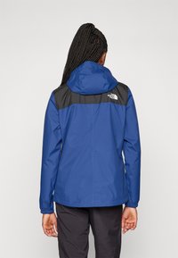Veste imperméable bleue avec un panneau noir sur les épaules, capuche réglable et poignets élastiques pour un ajustement sécurisé.