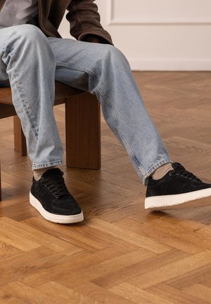 Personne portant un jean bleu clair, des baskets noires avec des semelles blanches, des chaussettes beiges et une veste marron, assise sur un banc en bois sur un sol en parquet.