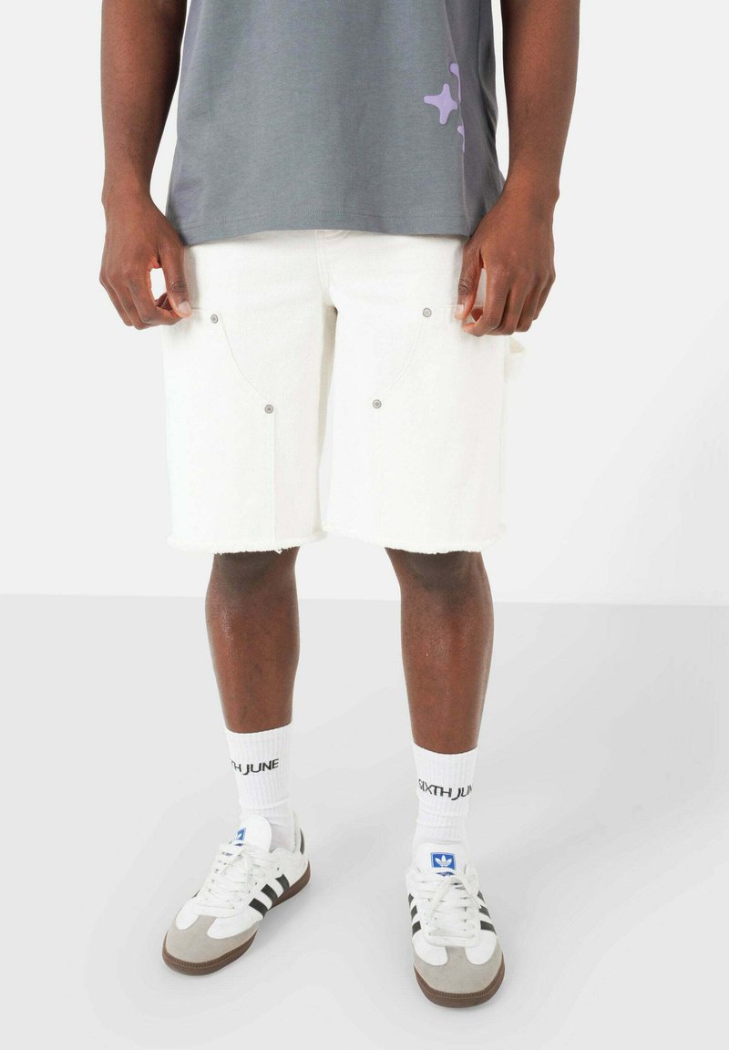 Pantaloni corti in denim bianco con orli sfilacciati e hardware argentato, abbinati a una maglietta grigia con una grafica viola. Indossati con calzini bianchi e sneakers.