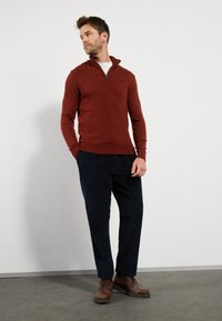 Dunkelroter Reißverschluss-Pullover mit hohem Kragen, gerippten Bündchen und Saum. Kombiniert mit marineblauen Cordhosen und braunen Schnürstiefeln.