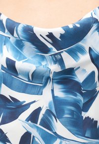 Μπλε και λευκό αφηρημένο μοτίβο φτερών σε λεία ύφανση, που πέφτει πάνω στον ώμο και το άνω μέρος του στήθους ενός ατόμου.