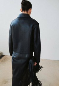 Długi czarny skórzany płaszcz z usztywnionym kołnierzem, o gładkiej fakturze i prostym kroju, w zestawie z czarną kopertówką z frędzlami.