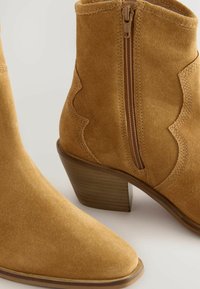 Botas de tobillo de ante color beige con punta afilada, cremallera lateral y diseño en forma de ondas. Presentan un tacón de madera apilada y una textura suave.