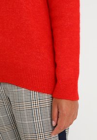 Maglione rosso lavorato a maglia con orlo e polsini a coste, abbinato a pantaloni a quadretti. Il maglione presenta una texture morbida e un colore vivace.