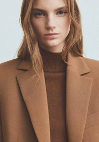 Blazer marrón con un diseño estructurado, combinado con un cuello de tortuga acanalado marrón oscuro. Textura suave, acentos mínimos y fondo neutro.