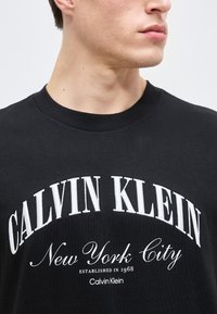 Κοντινό πλάνο ενός ατόμου που φοράει μαύρο t-shirt Calvin Klein με την επιγραφή "New York City" και "Established in 1968" σε λευκό χρώμα.