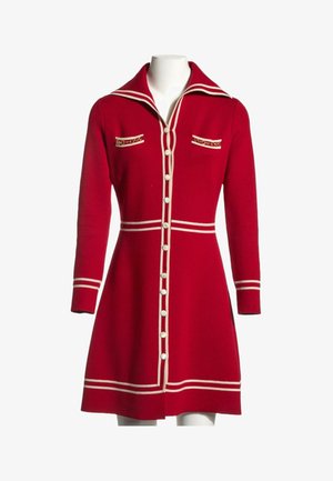 Robe rouge à manches longues boutonnée avec bordure blanche, grand col, deux poches poitrine avec détail chaîne et taille cintrée sur un mannequin.